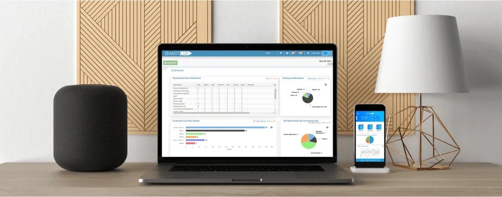 Laptop screen displaying the Qloud ERP system interface placed on a modern wooden desk alongside a smart speaker, smartphone, and decorative lamp.